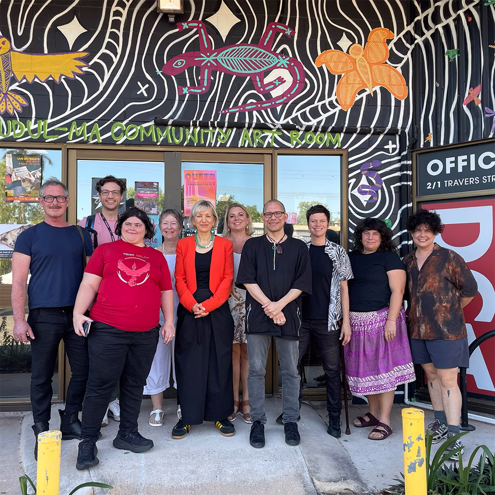 A group of ten people stand smiling below the entrance to a community centre. Framing the group is a black and white striped mural, featuring a painted purple lizard with teal linework.
