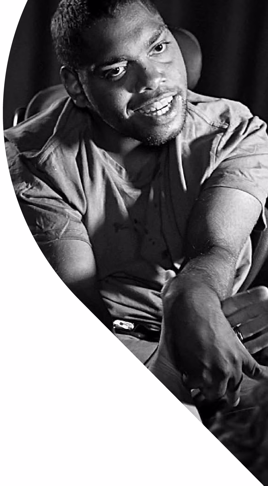 Photographed in black and white, a young man sitting in a power wheelchair, leaning forward and smiling. He has close cropped dark hair and stubble, and wears a short-sleeved shirt. 