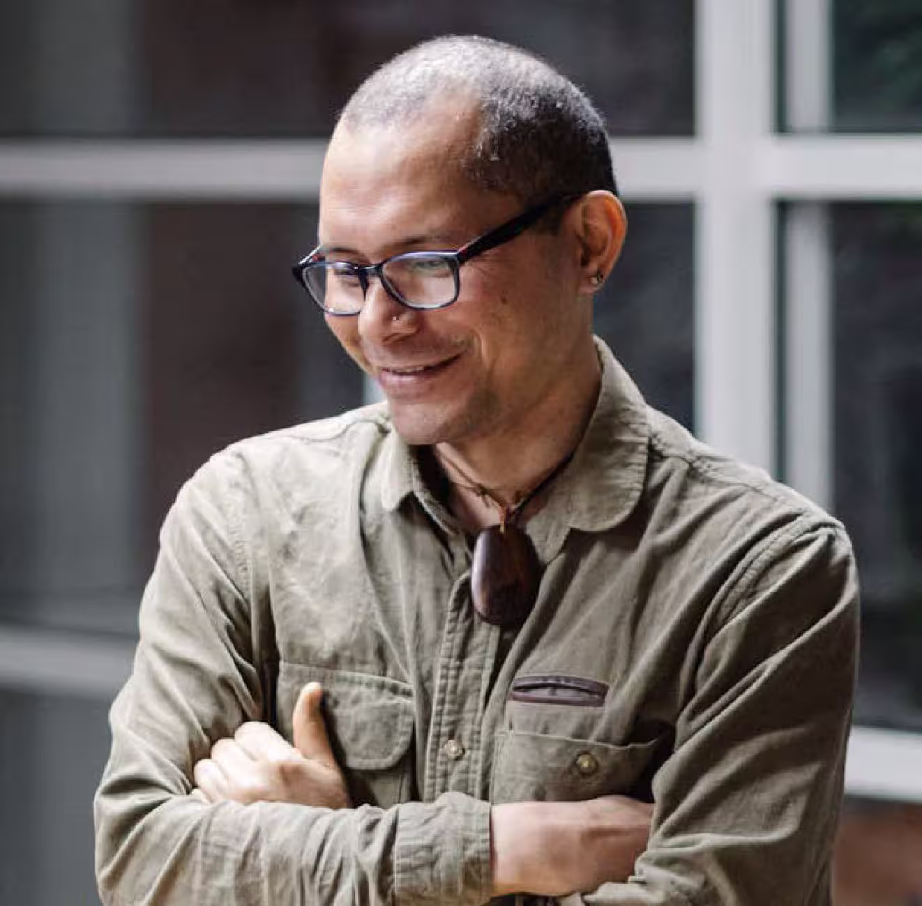 A man with short cropped dark hair, dark rimmed glasses and a khaki shirt smiles, his arms folded.