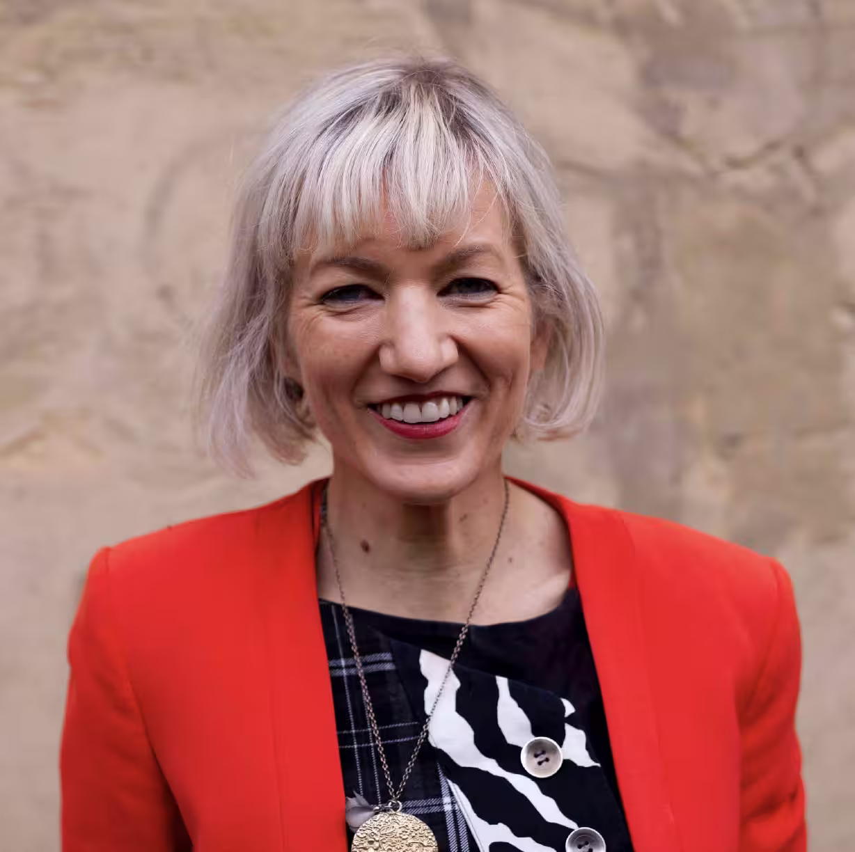 A woman, shown in profile, smiles widely before a blurred beige background. She has white-blonde hair cut just above her shoulders, and wears a red blazer over a black-and-white patterned blouse.
