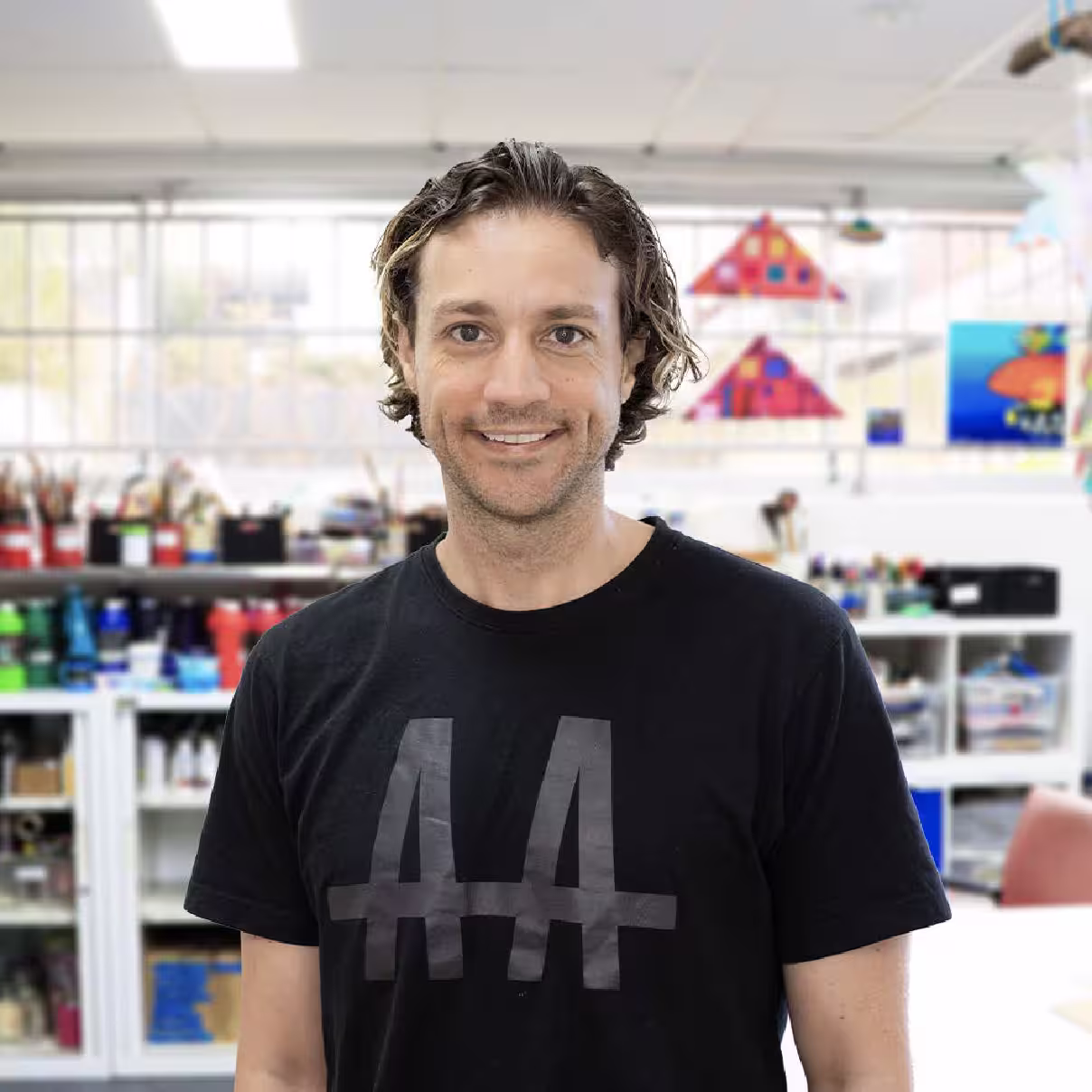 A young man with short brown wavy hair smiles towards us, standing in profile. He wears a black t-shirt with grey stylised text reading A A. Behind him in the distance are shelves of paints and art studio supplies.