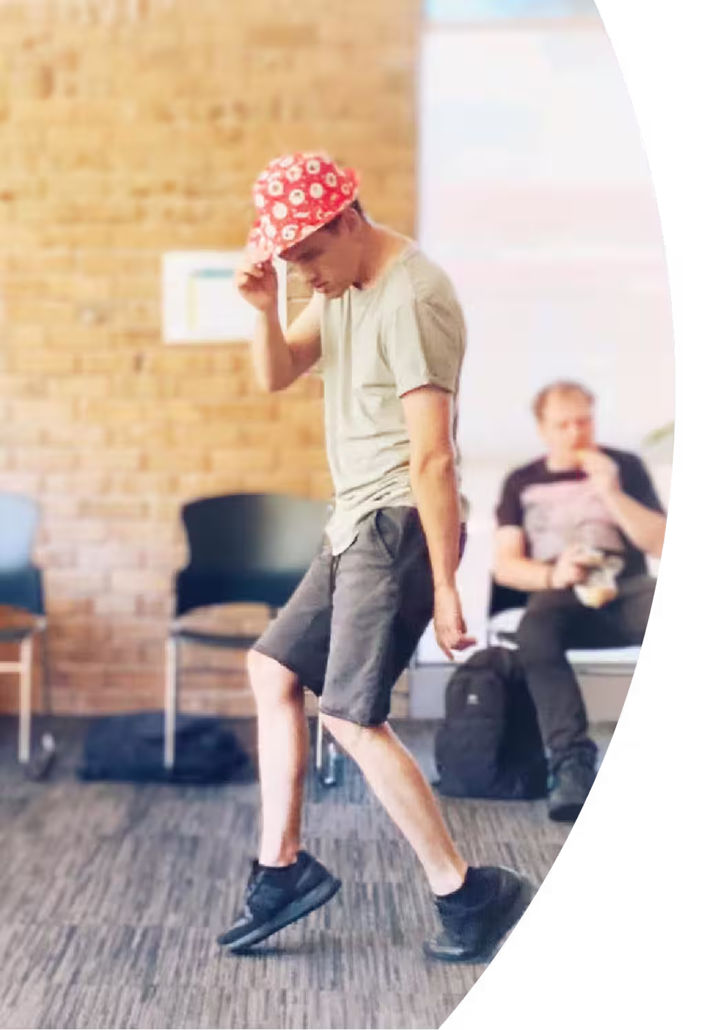 A young man dances, standing on tiptoes with one hand on a red and white patterned fedora hat. He wears a pale green t-shirt, grey shorts and black sneakers. 