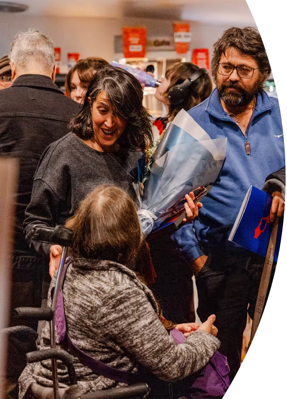 Three people talk in a crowded indoor place. A woman in a wheelchair turns her head to chat with a man and a woman standing to her left. The standing woman smiles as she speaks, holding a shiny silver-wrapped bouquet.